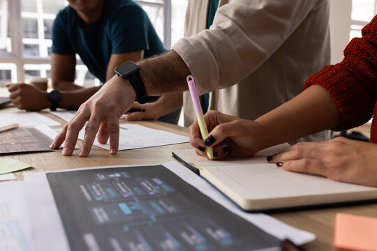 Coworkers reviewing mockup and noting in notebook at wooden table with pastel pen smartwatch