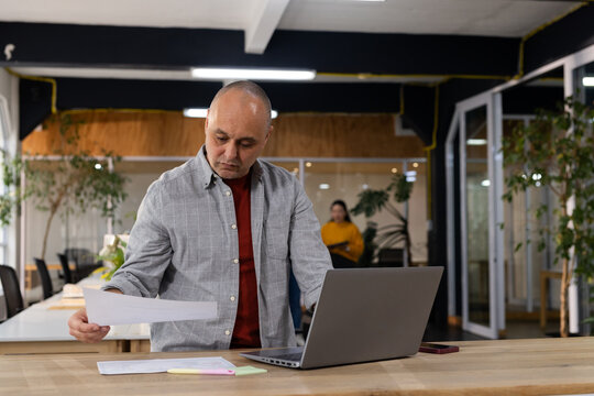 Diverse coworkers standing at wooden desk reviewing documents with laptop, smartphone in office