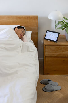 Asian woman sleeping on left side in bedroom with wooden headboard tablet lamp plant, copy space