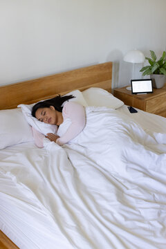 Asian woman sleeping in bed wearing light pink top under white duvet with tablet