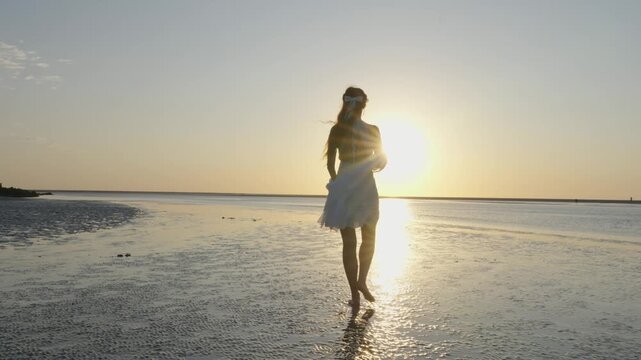 Woman in white dress walking barefoot across wet sand at sunset