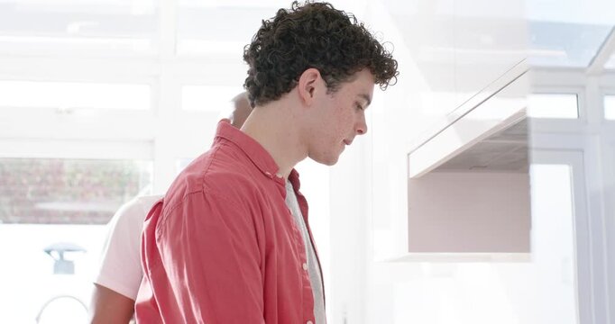 Diverse men cooking in kitchen, coral shirt man checking pan, grinding pepper to season, stirring