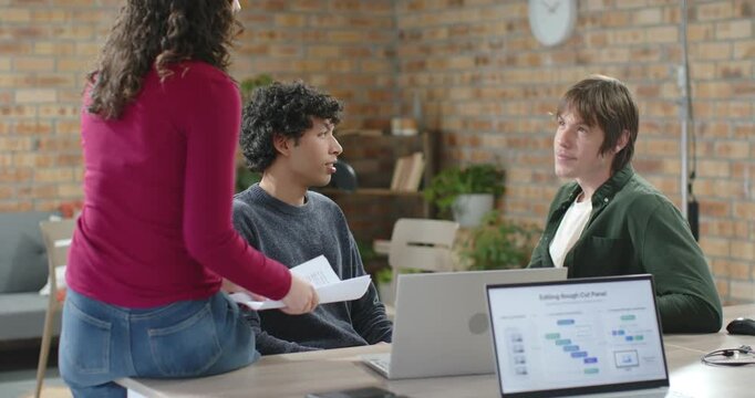 Three colleagues collaborating, woman approaching with printouts and guiding chart review on laptop