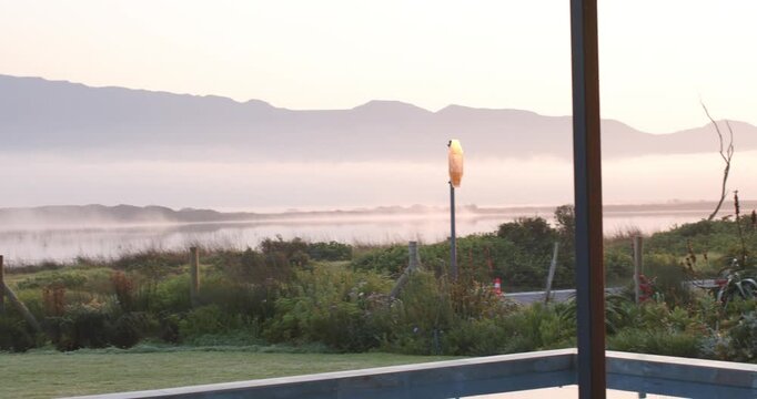 Camera is panning right through glass as warm sun is rising, revealing gate, fog and mountain