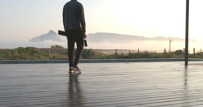 Sunrise drawing African American man in joggers walking across deck to sea, holding yoga mat
