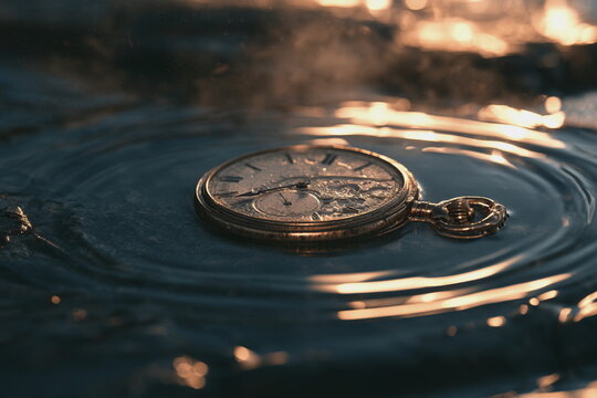 Antique pocket watch resting on dark rippling water in warm golden light, vintage timepiece, reflections, passage of time, nostalgia and timeless elegance.