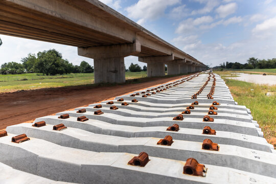 Rows of concrete railway sleepers with metal fastening shoulders awaiting rail installation beside an elevated concrete viaduct. 
