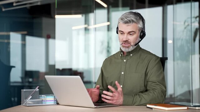 Gray-haired businessman using laptop and headset during online coaching session in office, guiding clients, providing professional advice, sharing knowledge, leading virtual training, communication.