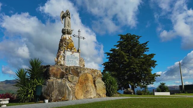 The historic Anjo de Portugal stone sculpture with a metal cross stands atop a rustic boulder pedestal under a bright cloudy sky.
