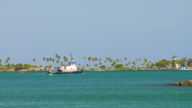 Tugboat travels along the San Juan bay as it slowly it moves along the turquoise waters of the Caribbean during a sunny day. Camera movement right