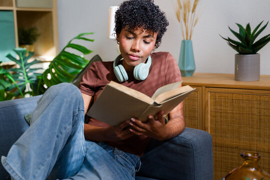 African American woman reading book on sofa with headphones