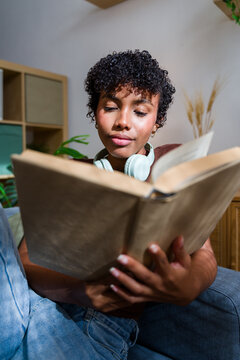 African American woman reading book indoors with headphones on sofa