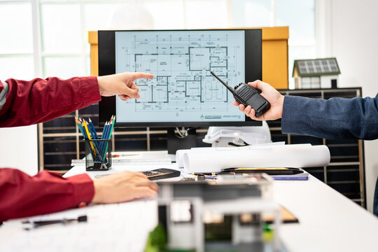 Architects or engineers collaborate over a building floor plan on a computer, with drafting tools, blueprints, a scale model, and a handheld radio in a bright office.