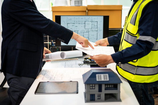 Architect and construction worker review house plans at a desk with blueprints, tablet, computer drawing, and a small home model.