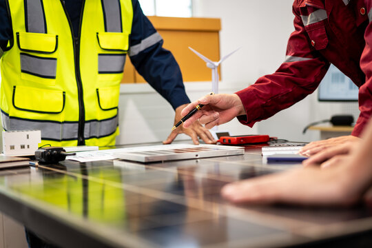 Engineers in safety clothing discuss solar panel plans at a desk, with wind turbine model and tools in a bright renewable energy office.