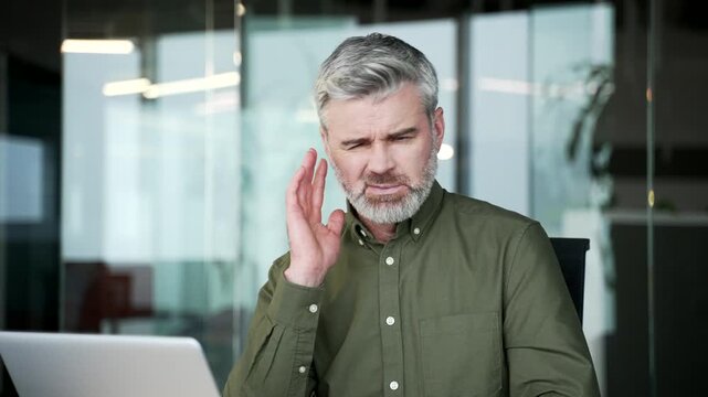 Close-up of businessman suffering from ear pain. Sick mature man touching ear with fingers, feeling discomfort and ache caused by infection, otitis or ear disease while sitting at office workplace.