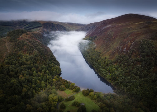 Aerial view of Glendalough Upper Lake surrounded by lush green forests, steep hills, and low-hanging clouds in the Wicklow Mountains, Glendalough, County Wicklow, Ireland.