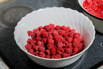 A white ribbed bowl filled with ripe red raspberries on a modern induction cooktop ready for homemade jam. © Konstantin