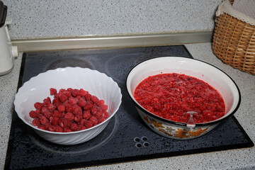 Fresh red raspberries in a white bowl next to crushed berry mass in an enamel basin on a kitchen cooktop. © Konstantin