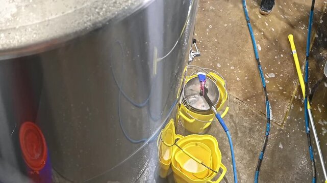 A high-angle shot of a stainless steel brewing tank during cleaning at a craft brewery, with yellow buckets and hoses on the wet floor.