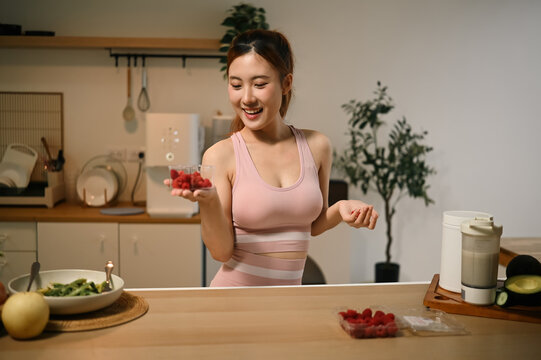 Young Asian Woman in Sportswear Holding Bowl of Fresh Raspberries in Modern Kitchen for Healthy Lifestyle