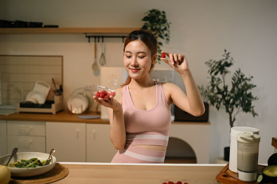 Young Asian Woman in Sportswear Holding Raspberries in Modern Kitchen Enjoying Healthy Antioxidant Rich Snack