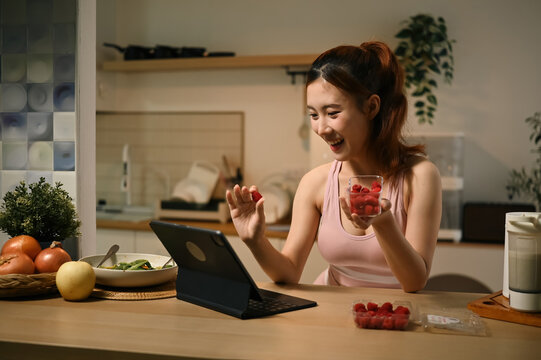 Young Asian Woman Eating Raspberries While Using Tablet in Kitchen, Healthy Lifestyle and Antioxidant Benefits