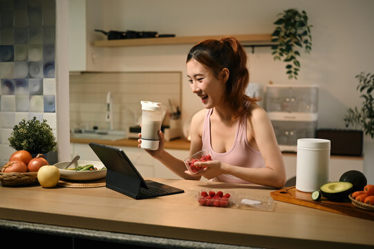 Young Asian Woman Holding Raspberries and Protein Shake in Kitchen While Browsing Healthy Nutrition Tips on Tablet