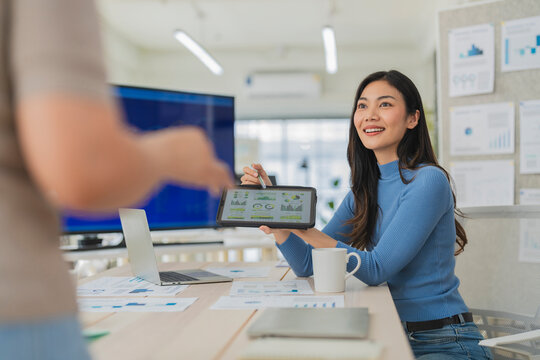 Smiling young Asian businesswoman presenting data dashboard on digital tablet to colleague during office meeting