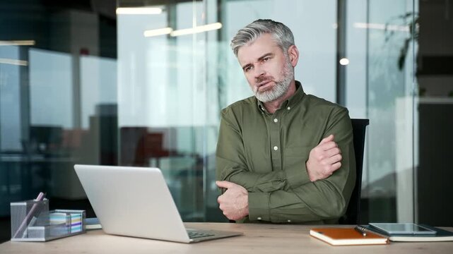 Gray-haired businessman sitting at office desk suffering from elbow pain. Mature man touching massaging joint, rubbing sore muscles and feeling discomfort or tension while working at modern workplace.