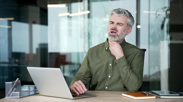 Sick gray-haired businessman sitting at office desk suffering from sore throat. Mature man touching and massaging neck with hand, feeling pain and discomfort while working at modern workplace.