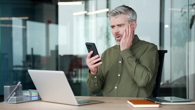 Mature gray-haired businessman sitting at desk in office, holding smartphone and reading bad news, shocked and disappointed by unexpected message, frustration, disbelief, stressful situation at work.