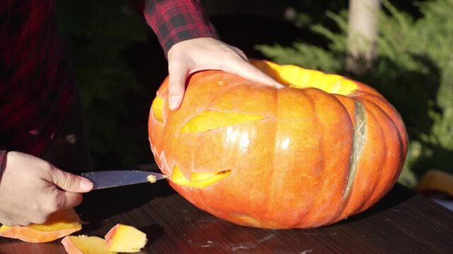 Close up of hands carving scary grinning mouth pushing out ripe squash flesh using kitchen knife in golden hour, authentic autumn holiday craft