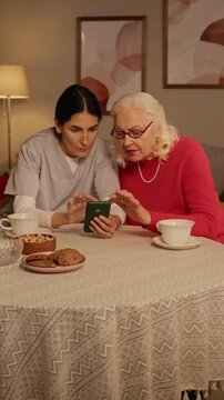 Vertical video of female caregiver showing smartphone to senior Caucasian woman at table. Clip suitable for senior healthcare services, assisted living ads, or elderly digital literacy projects
