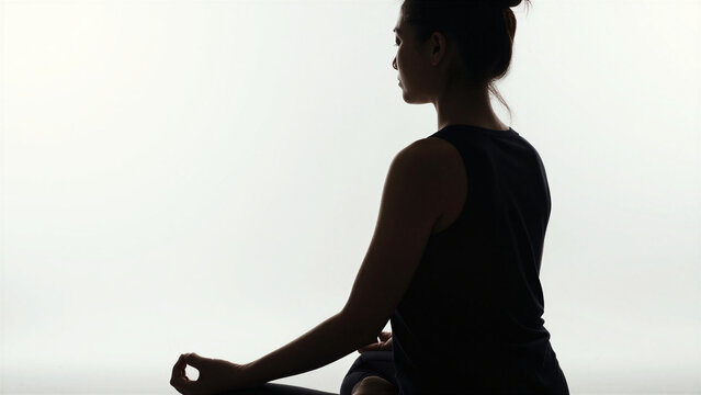 Profile silhouette of woman meditating in zazen pose with bright morning sunlight background, copy space on the left, diagonal back view.