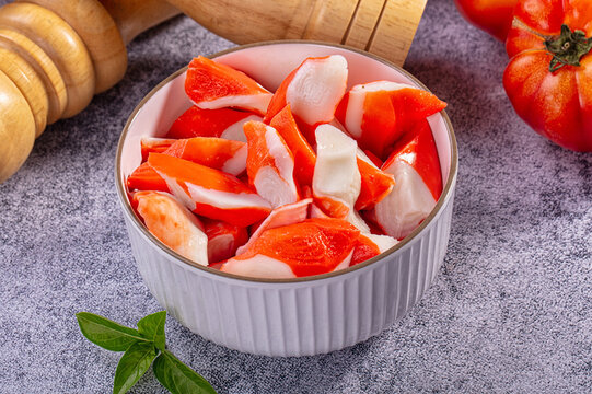 Grey stone table with white ceramic bowl of chopped surimi crab sticks, wooden pepper mills and ripe tomato, fresh ingredient concept