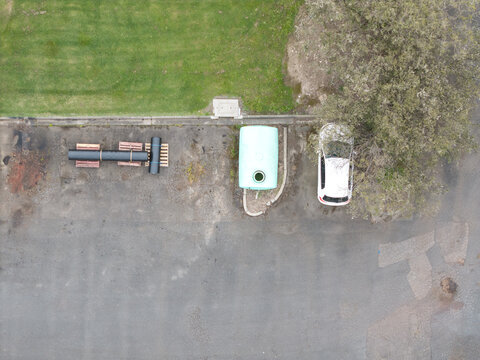 Aerial view of Bayswater North Factories featuring a large light-blue industrial tank, a white car parked near a tree, and black pipes on pallets on the asphalt, Melbourne, Victoria, Australia.