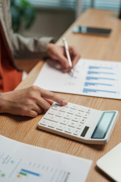 Close-up of a professional using white calculator over printed financial charts at a wooden desk. Budget figures and business reports are reviewed together during accounting work.