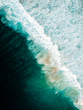 Aerial view of turquoise ocean waves crashing onto the sandy shore of Pantai Watu Bella in Lamboya, East Nusa Tenggara, Indonesia.