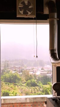 Vertical view from a roastery room window with industrial exhaust pipes, revealing a scenic rural village and mountain landscape