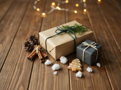 Two christmas gift boxes wrapped in brown and grey paper with pine sprigs, cinnamon, pinecones, and a gingerbread tree on a dark wooden background
