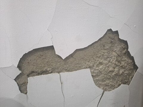 Close-up of cracked and peeling white paint on wall exposing rough gray concrete plaster underneath, damaged surface texture, building maintenance concept