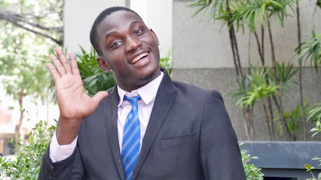 20s young adult black African businessman waving hello to camera in outdoor urban office scene, friendly smile and professional suit