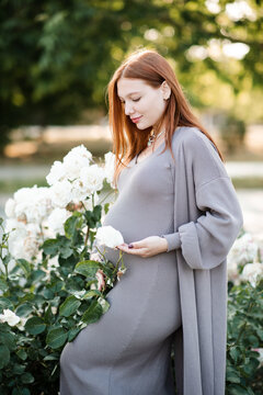 Stylish smiling pregnant woman wearing maternity clothes with rose flowers in garden outdoors. Prenatal lifestyle. Spring season. Motherhood.