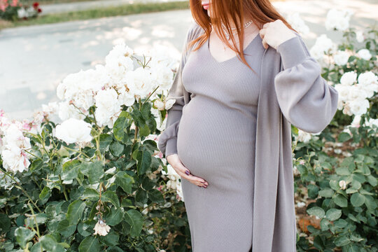 Pregnant woman wearing maternity clothes with rose flowers holding tummy in garden outdoors close up. Prenatal lifestyle.