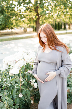 Smiling cute pregnant woman 24 - 29 year old wearing maternity clothes with rose flowers in garden outdoors. Prenatal lifestyle.