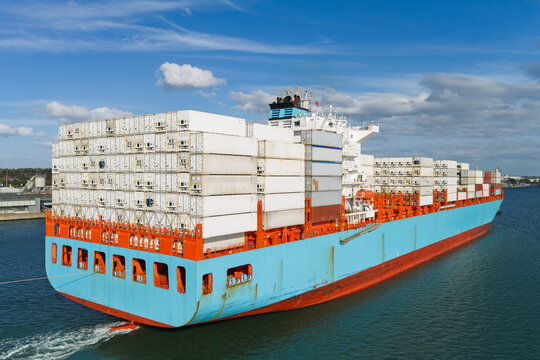 Aerial view of a large container ship loaded with white shipping containers sailing through calm coastal waters under a blue sky with scattered clouds in the United Kingdom.