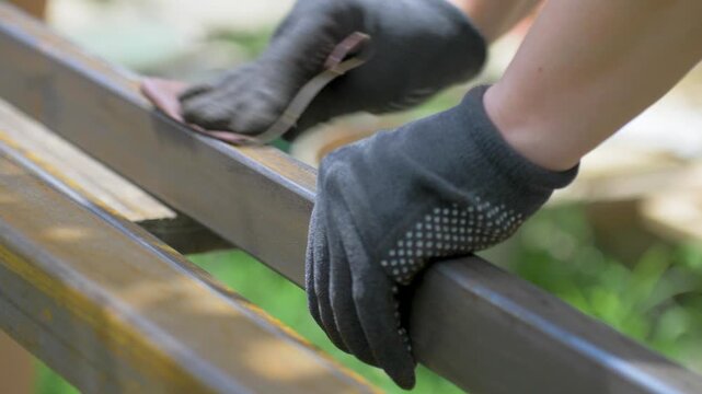 Gloved hands hold coarse sandpaper over old rusted iron beam. Direct pressure polishes metal surface on wooden support bench. Outdoor manual restoration revives worn steel construction material