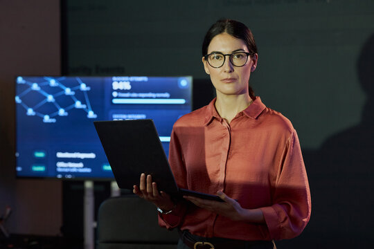 Portrait of Caucasian young adult woman holding laptop in cybersecurity office with data screen background. Visual concept for IT security, software development marketing