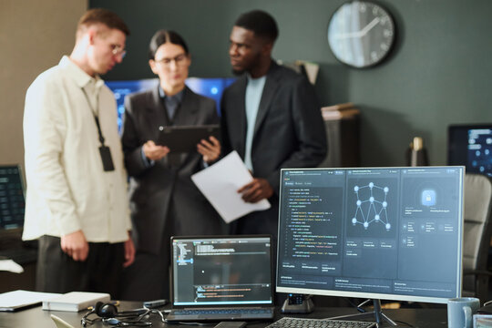 Multiethnic young adult cybersecurity team analyzing network data in office while computer screens show security dashboard. Ideal for IT safety, tech business, data protection projects